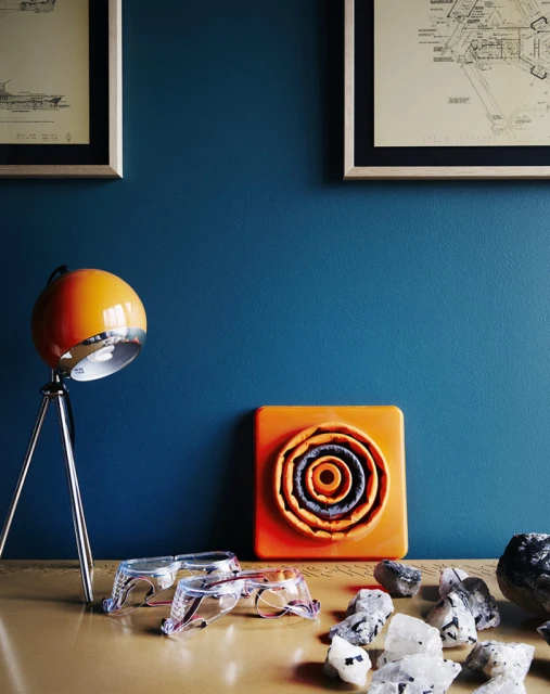 Still life featuring an orange lamp, a square orange concentric design object, glasses, quartz crystals on a beige surface against a blue wall.