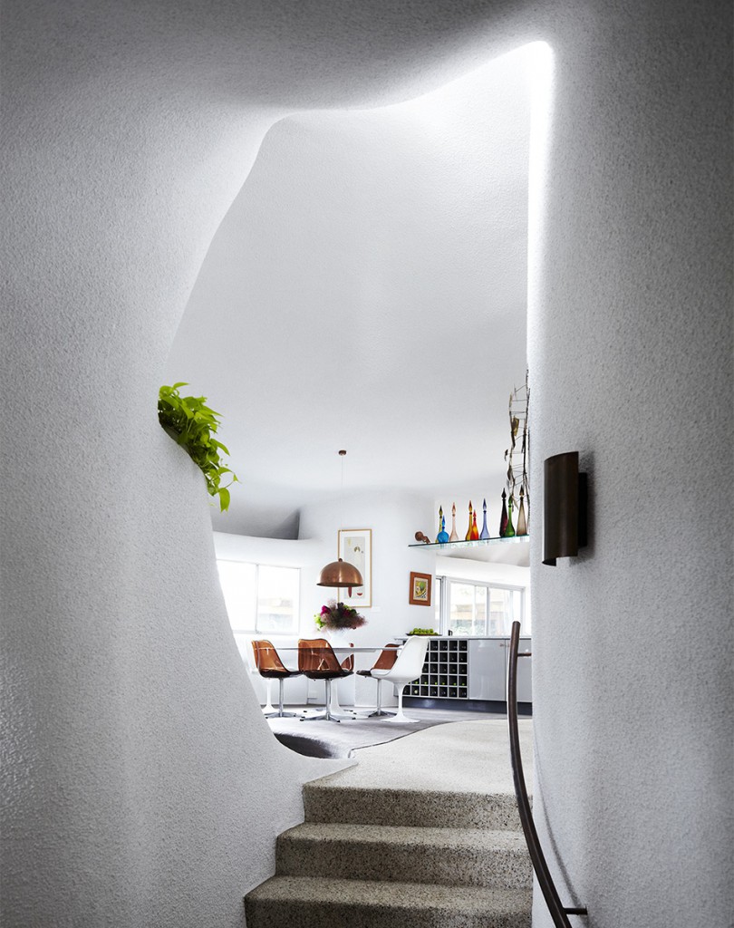 Modern architecture showcasing an indoor staircase leading to a dining area with white walls, a unique organic shaped entrance, and plants.