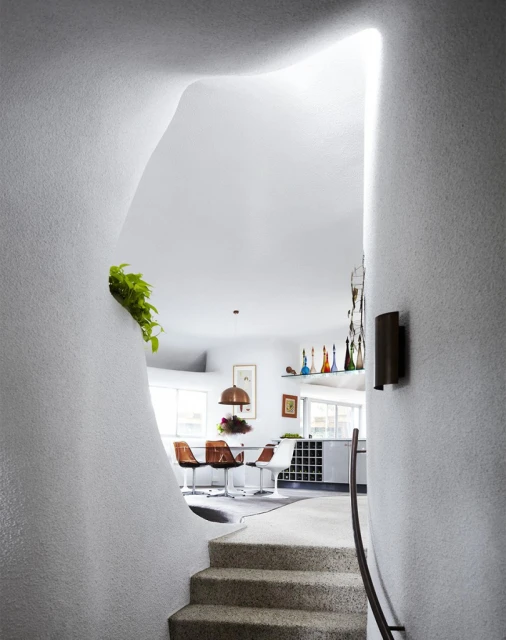 Modern architecture showcasing an indoor staircase leading to a dining area with white walls, a unique organic shaped entrance, and plants.
