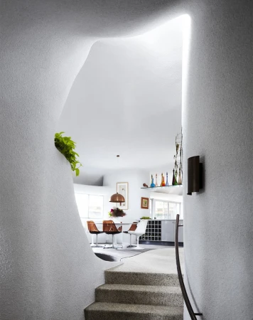 Modern architecture showcasing an indoor staircase leading to a dining area with white walls, a unique organic shaped entrance, and plants.