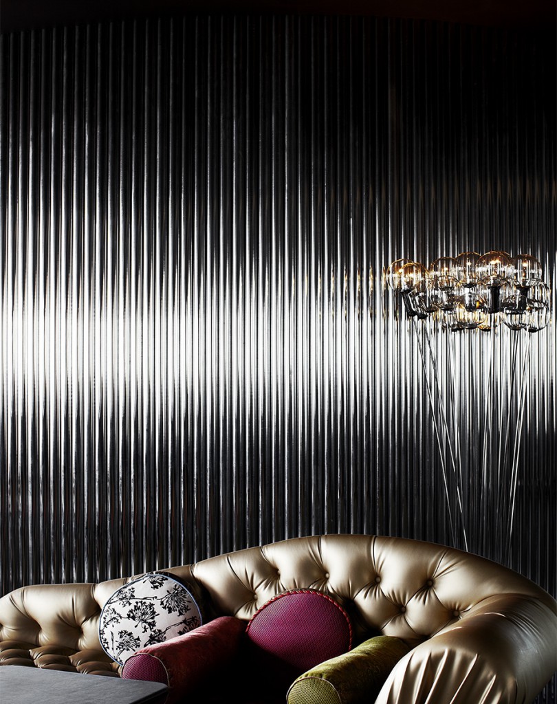 Interior design of a living room with a tufted gold sofa and decorative throw pillows against a black, corrugated wall with a modern chandelier.