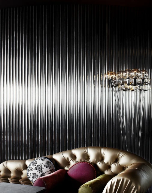 Interior design of a living room with a tufted gold sofa and decorative throw pillows against a black, corrugated wall with a modern chandelier.
