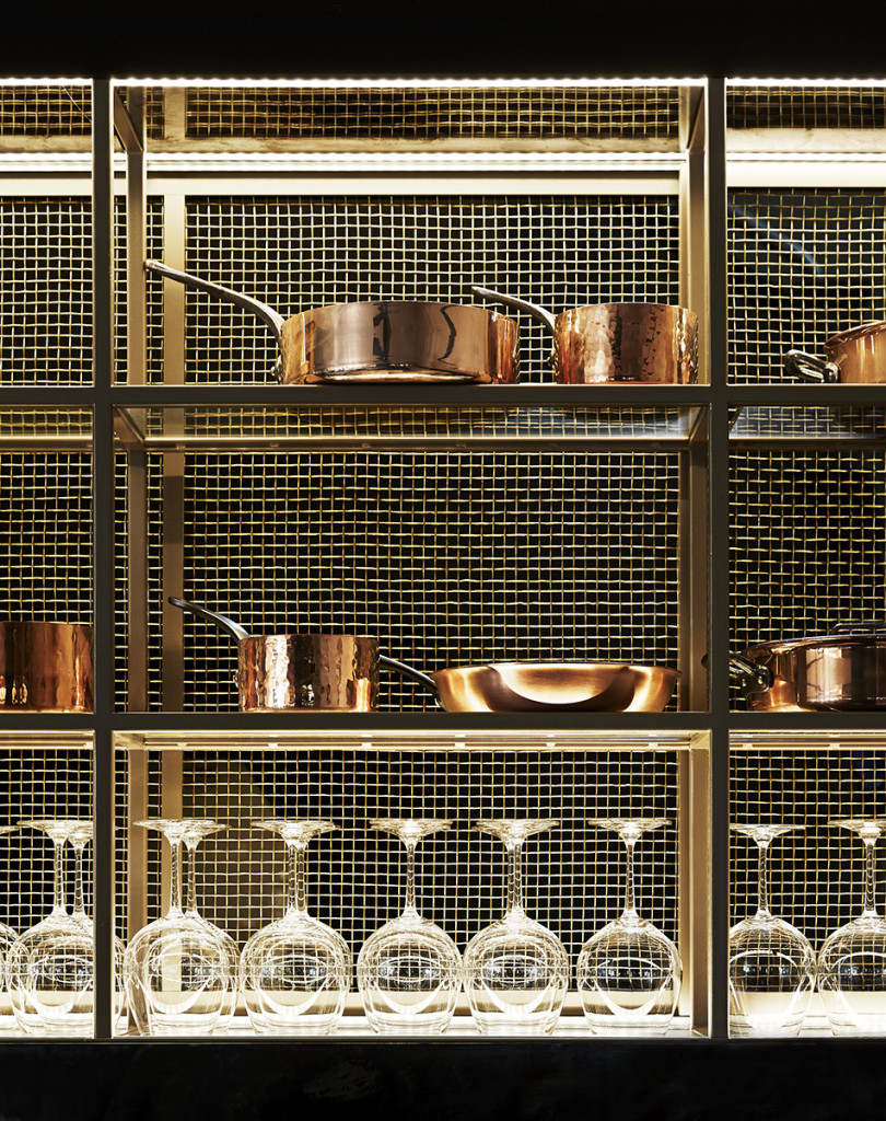 A well-lit kitchen display featuring copper cookware and wine glasses arranged on a gold metal shelving unit with a wire mesh backing.