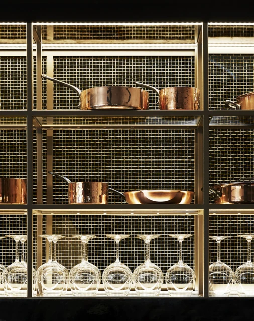 A well-lit kitchen display featuring copper cookware and wine glasses arranged on a gold metal shelving unit with a wire mesh backing.