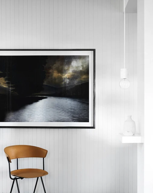Framed art print with dark river landscape hangs on a vertically paneled wall, next to a tan chair and a decorative white vase.