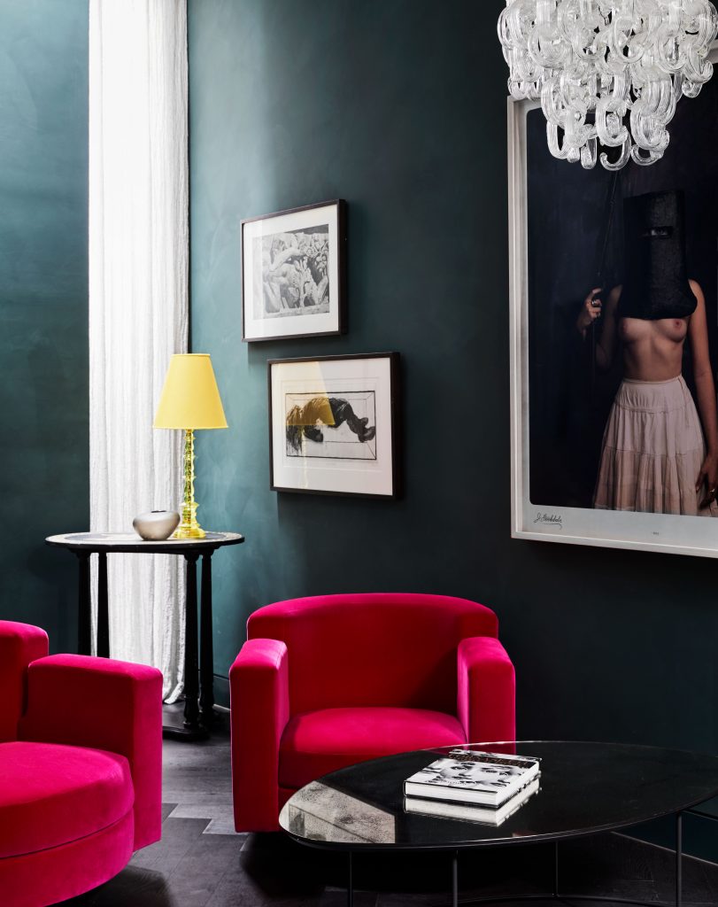 Eclectic living room interior featuring two pink velvet armchairs, black coffee table, lamp, and artwork against a dark teal wall.