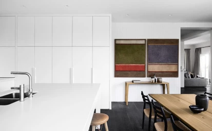 Modern minimalist kitchen interior with white floor-to-ceiling cabinetry, wooden dining table, and two abstract artworks on the wall.
