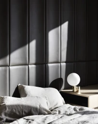 White linen bedding and pillows in front of a dark upholstered panel wall with a modern globe bedside lamp.