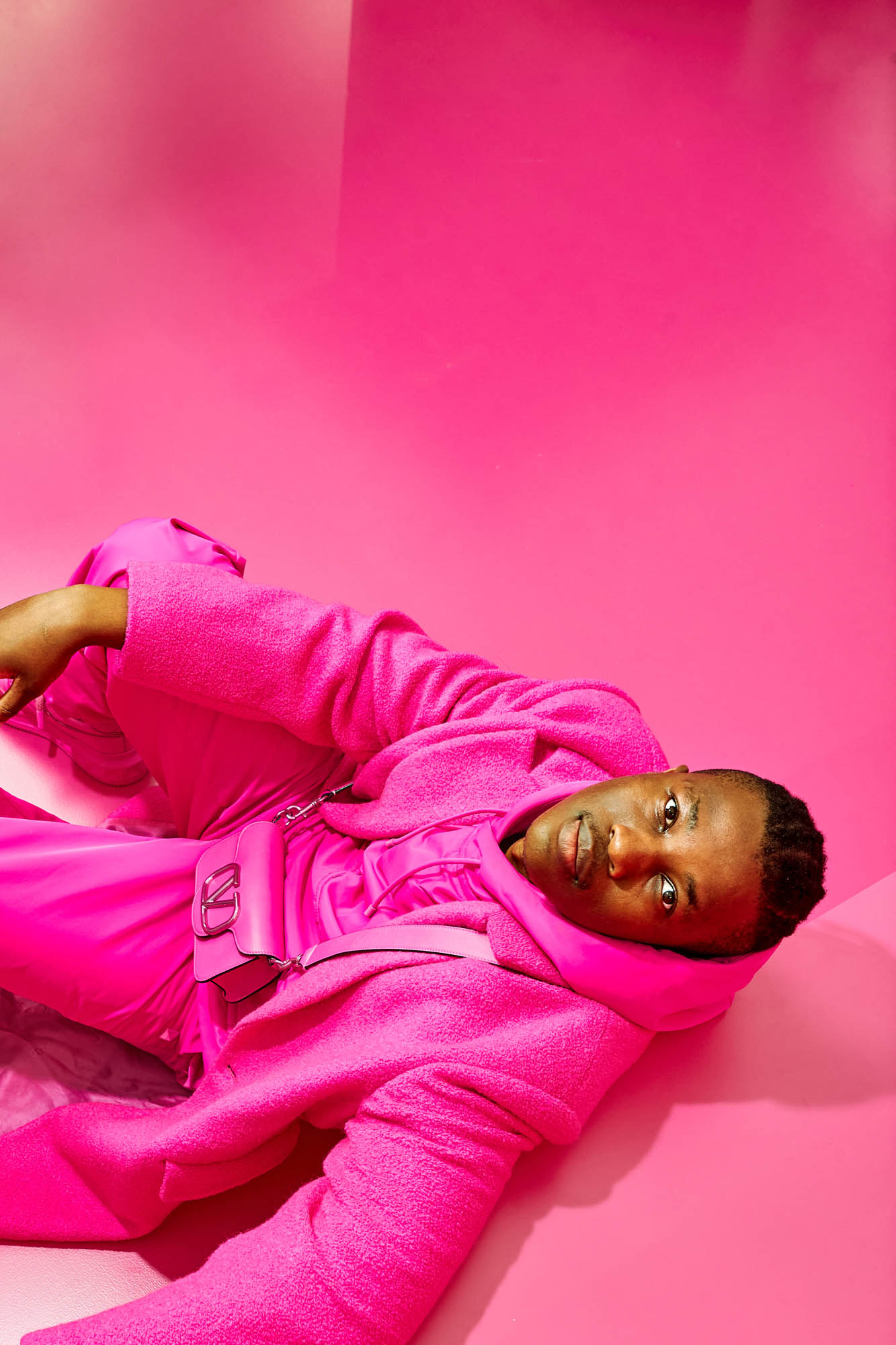 Fashion portrait of an adult male laying down in a vibrant pink matching hooded jacket, pants, and crossbody bag against a pink backdrop.