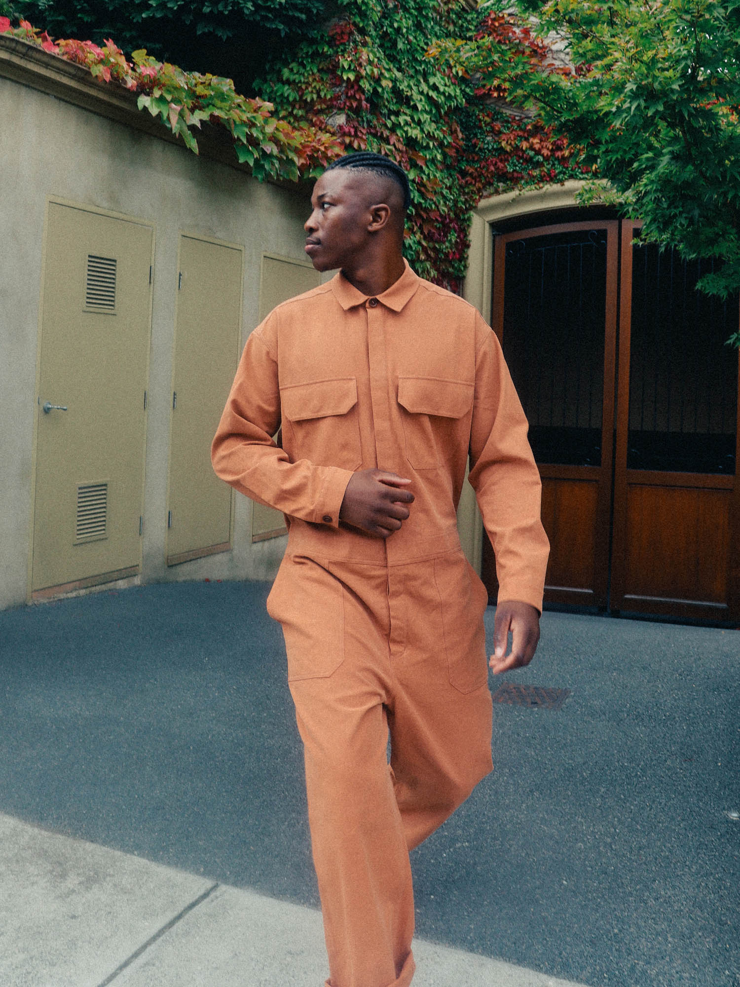Man in orange jumpsuit walking in urban setting near building with vine covered walls and closed doors, wearing long sleeves.