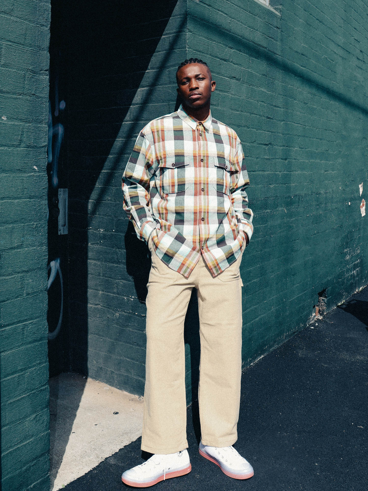 A young African-American man poses against a teal brick wall wearing a plaid shirt and khaki pants, bathed in sunlight. Street scene.