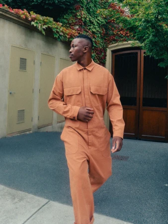 Black man wearing a rust orange jumpsuit walking past a concrete wall and dark wooden double doors.