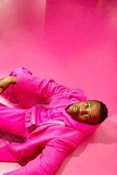 Black man wearing vibrant pink jacket and trousers lying against a matching pink studio backdrop