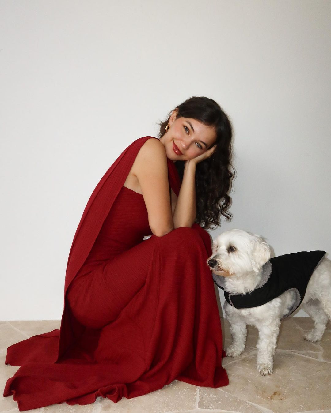 A woman in a red dress poses with a white dog wearing a black vest against a plain white background in a brightly lit setting.