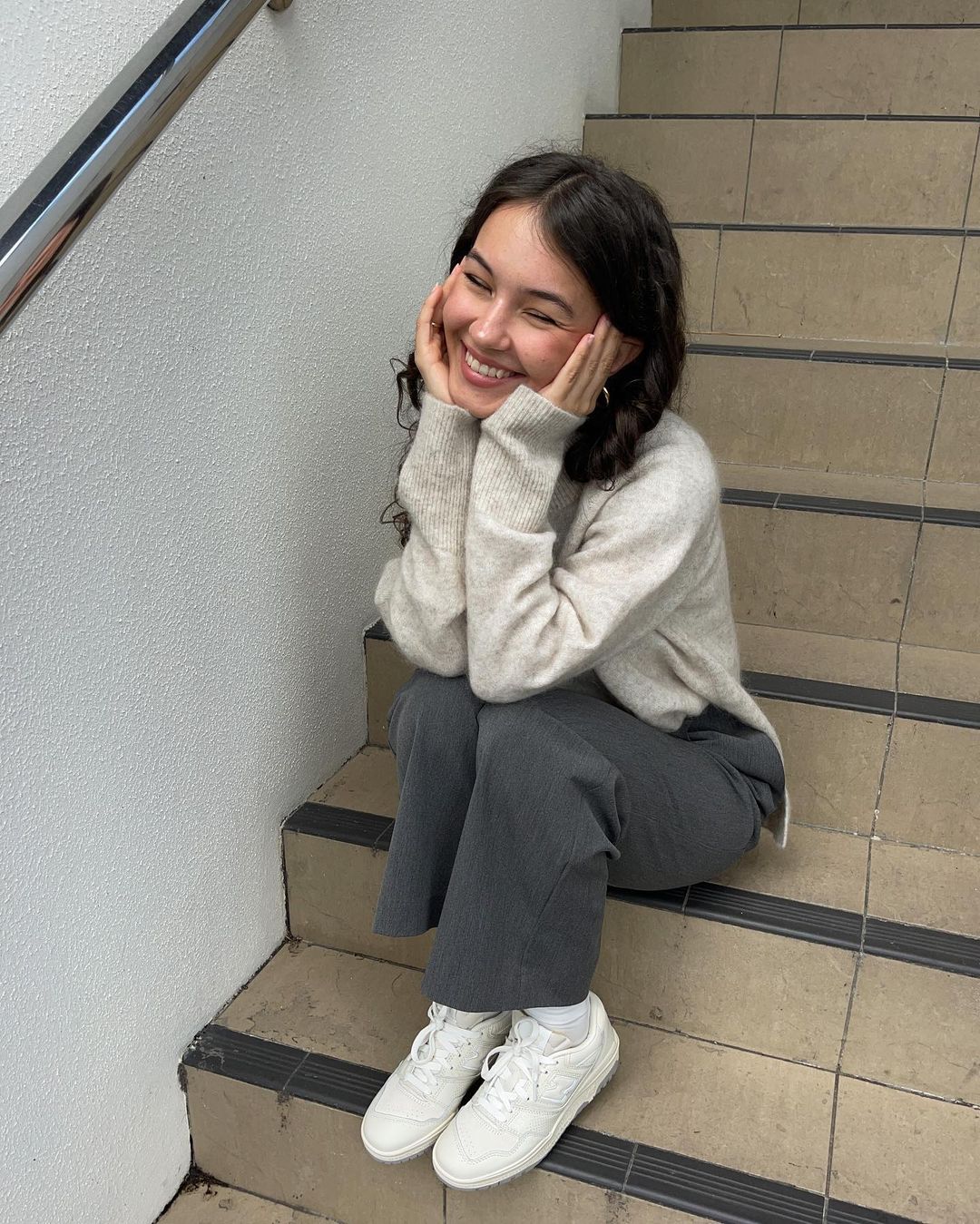 A young woman with dark hair and a warm smile sits on steps, her hands gently touching her cheeks in a relaxed and happy pose.