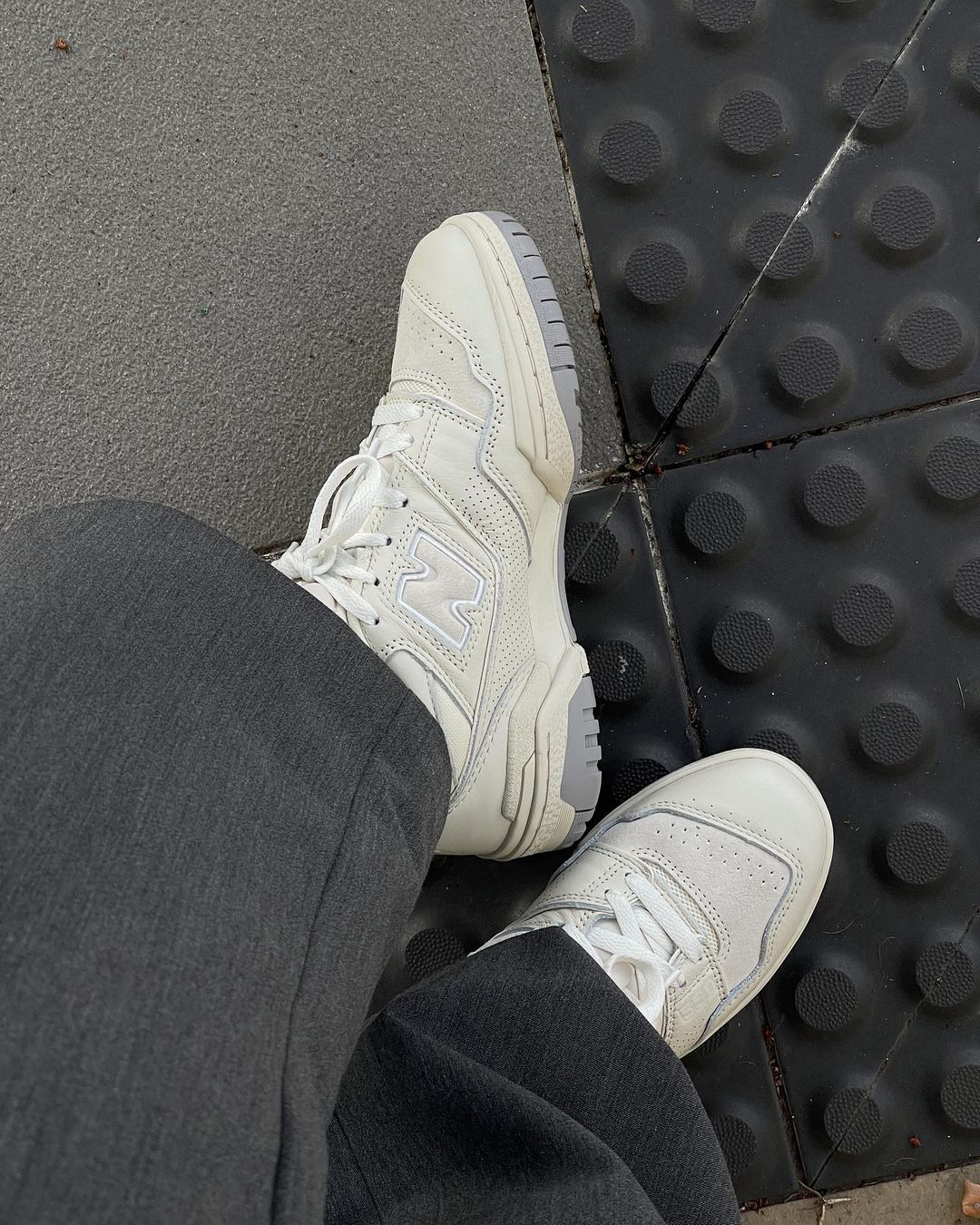 Overhead shot of someone wearing New Balance 550 sneakers in off-white and grey, paired with gray pants on asphalt.