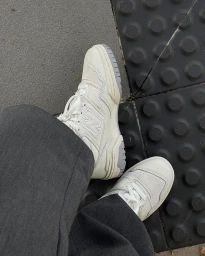 Close-up of a person wearing white sneakers and dark trousers standing on concrete and textured tactile paving tiles