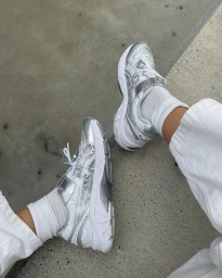 Close-up of white and silver sneakers paired with white socks and loose white trousers against a concrete surface.
