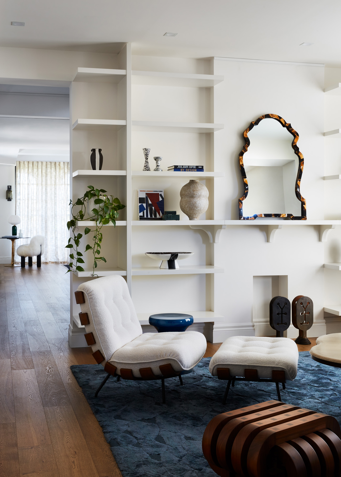 Interior shot showcases a cream-colored lounge chair and ottoman on a blue patterned rug, complemented by modern decor and a white bookshelf.