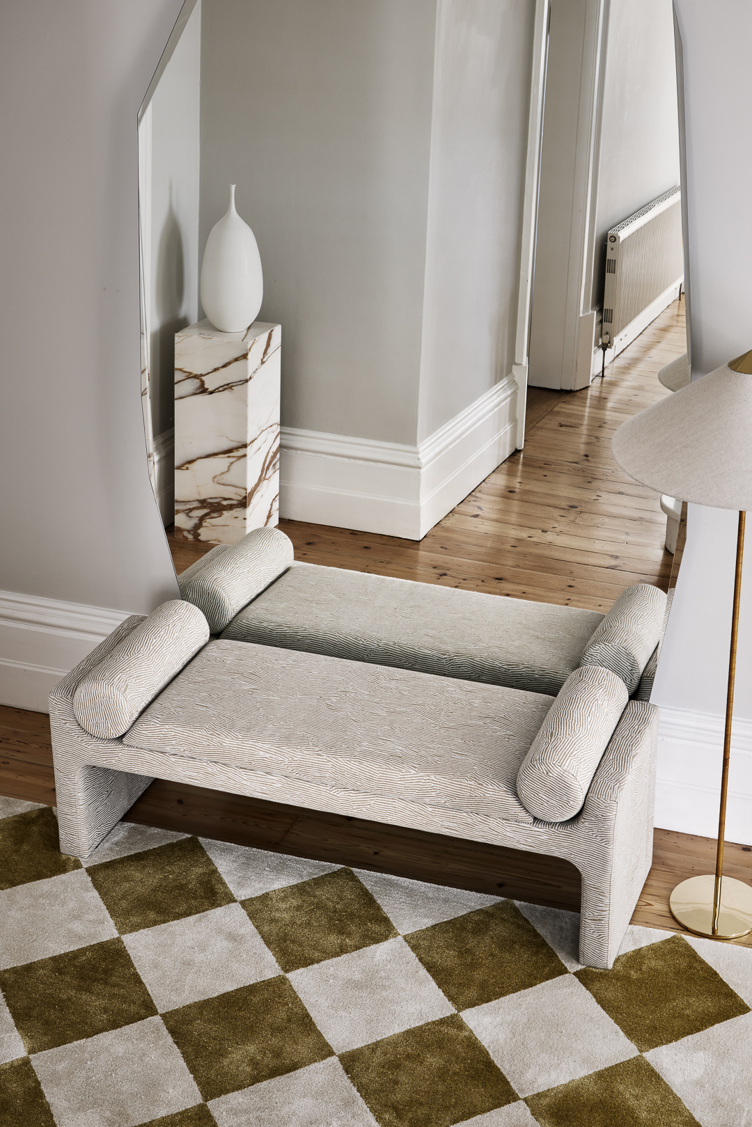 Interior design: Modern bench with bolster pillows on a checkered rug. Decorative vase and large mirror are in the background.