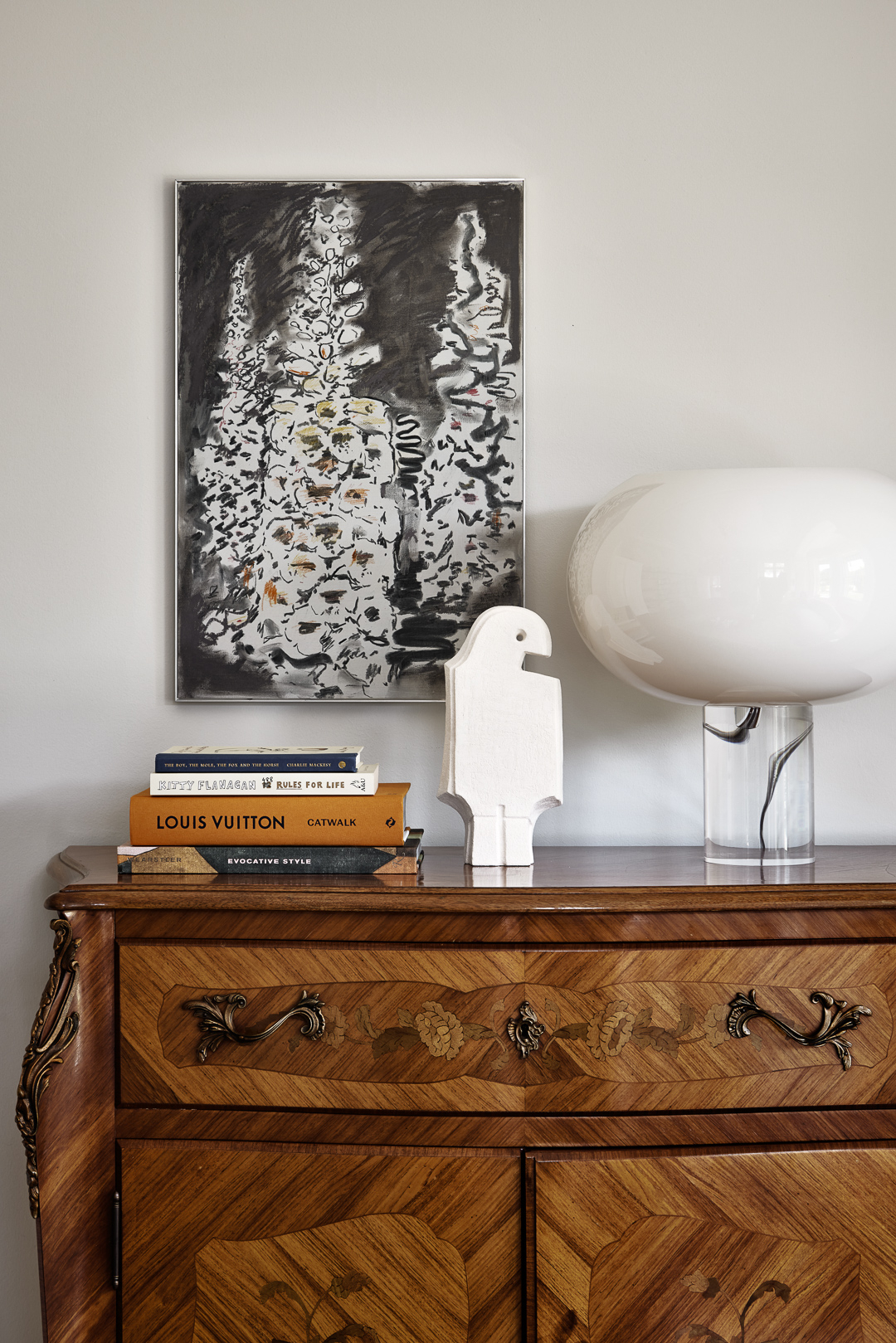 Interior view of a wooden dresser with books, artwork, a lamp, and a decorative figurine in an indoor setting with neutral tones.