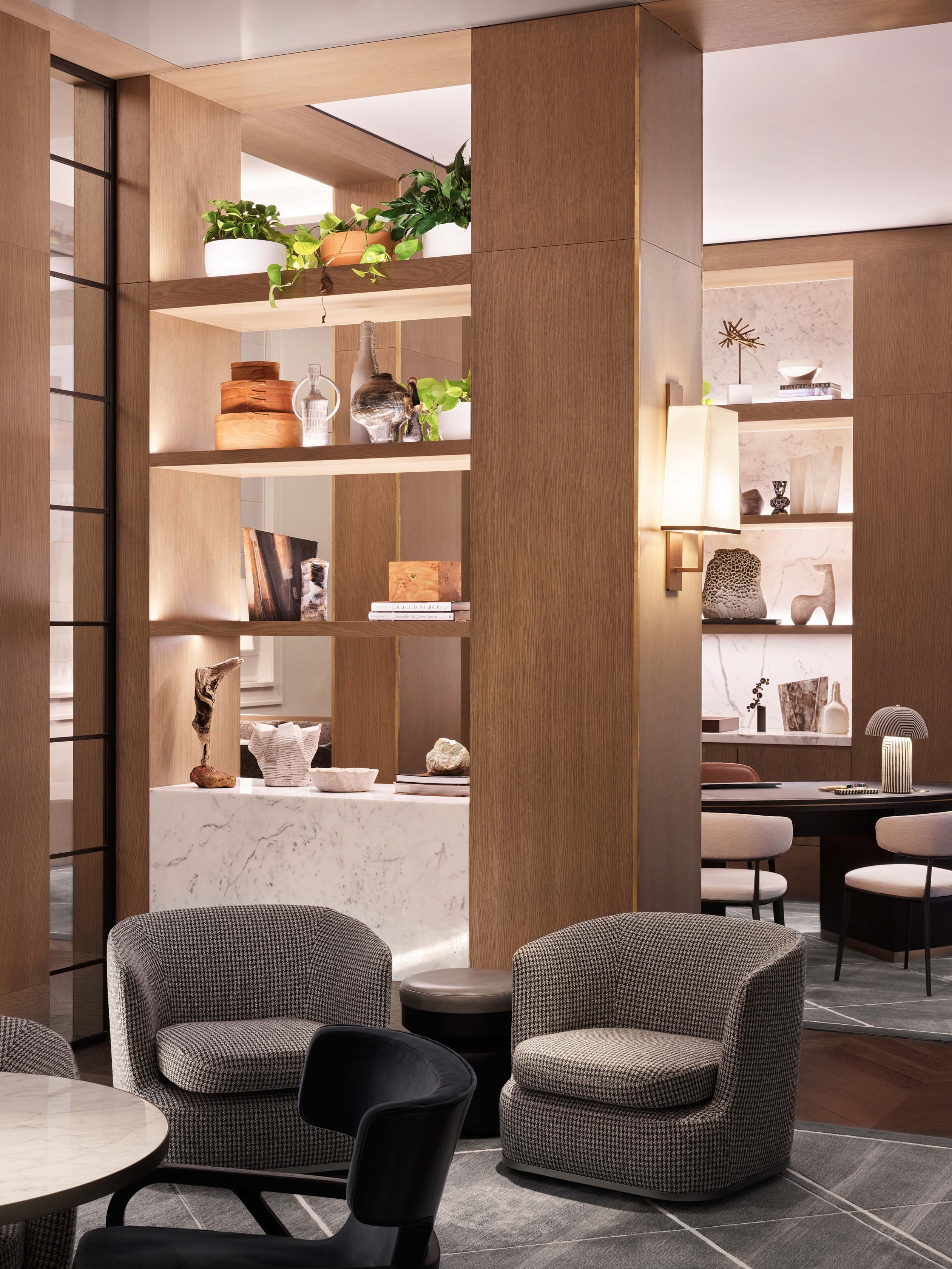 Interior view of a hotel lobby with several armchairs around small tables, a wooden shelving unit, wall sconces, and neutral color palette.