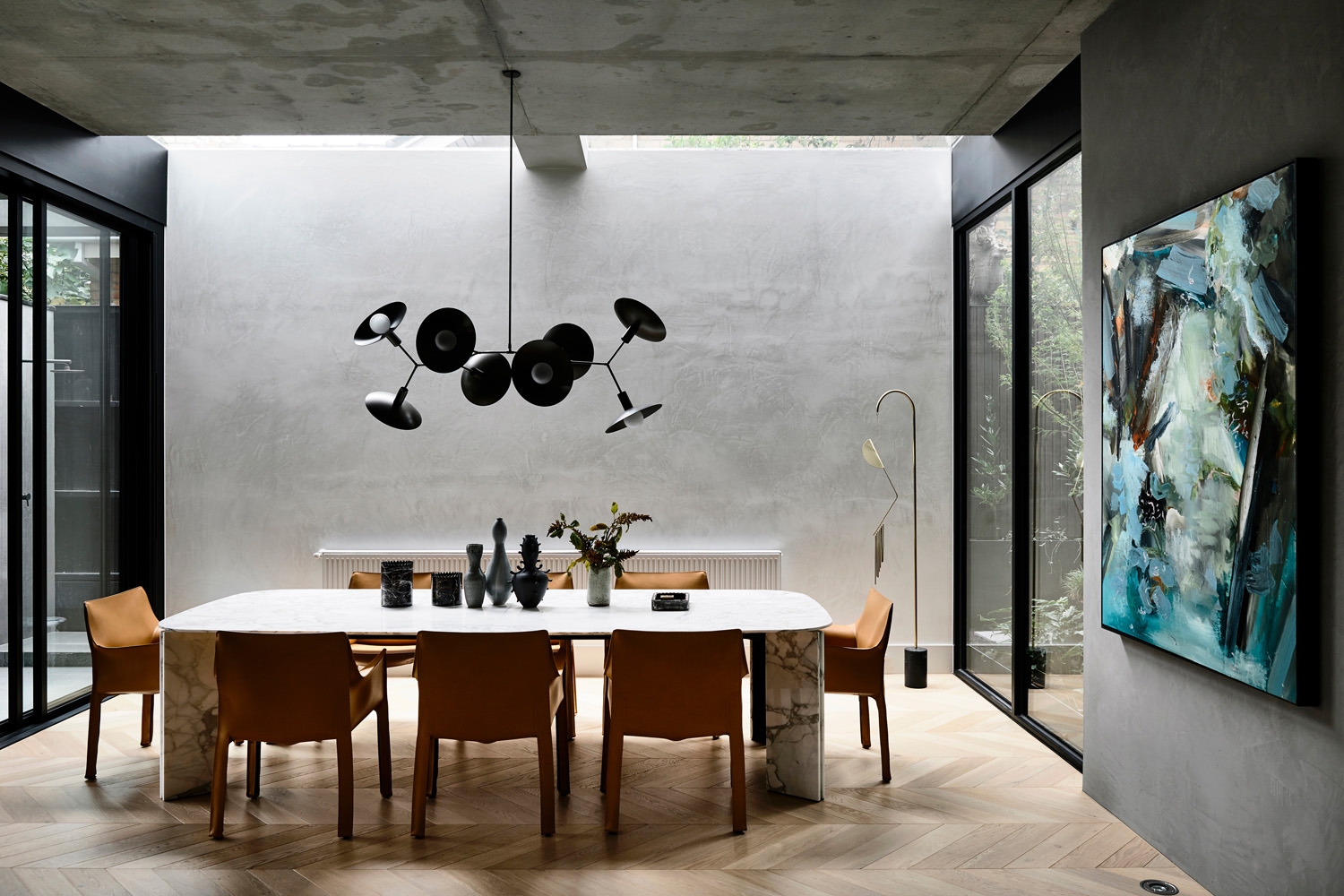A modern dining room features a marble-topped table surrounded by tan leather chairs, illuminated by a unique black chandelier.