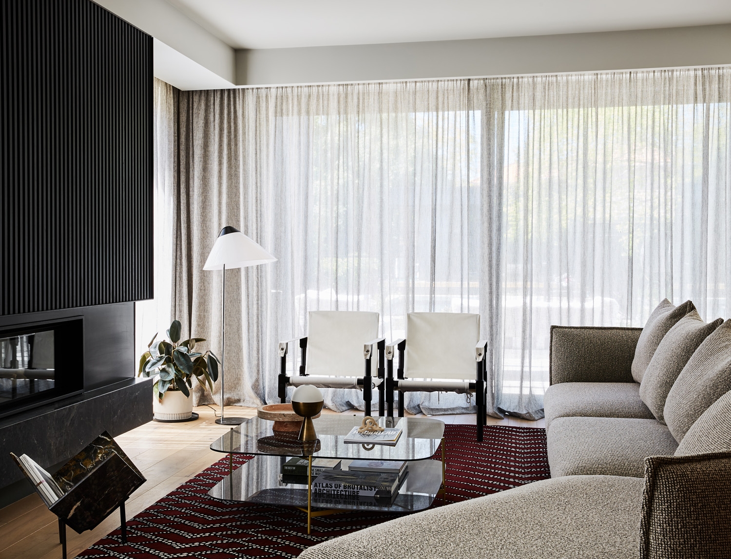Living room interior featuring a grey sofa, modern furniture, glass coffee table, area rug, sheer curtains and natural light.