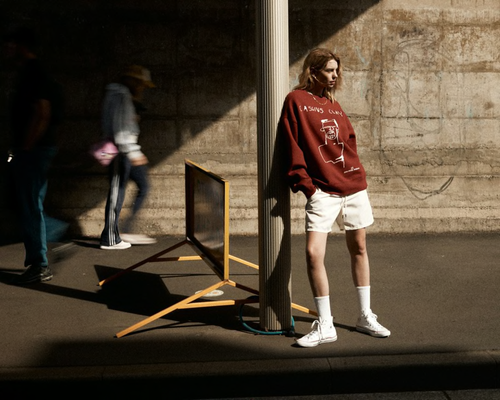 A young adult poses near a pillar in a burgundy sweatshirt, white shorts and sneakers with a cement wall and pedestrians in the background.