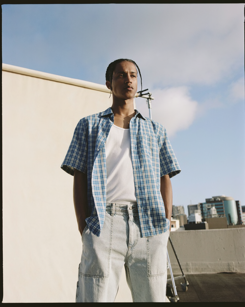 An adult male standing outside, wearing a blue and white checkered shirt over a white tank top and light blue pants, under a blue sky.