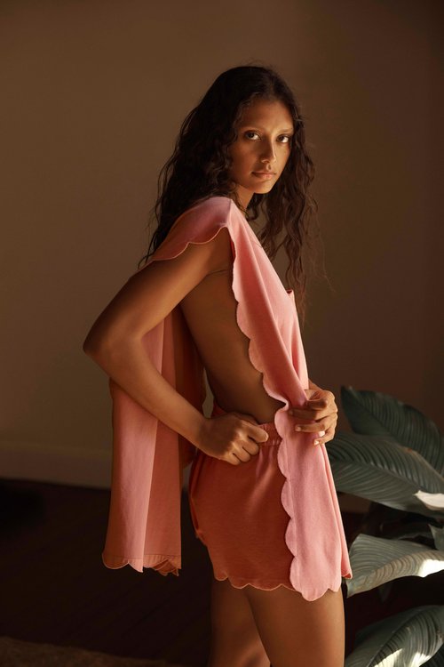 A woman wearing a pink scalloped-edge outfit stands indoors, showcasing her back in a natural light setting with plant background.