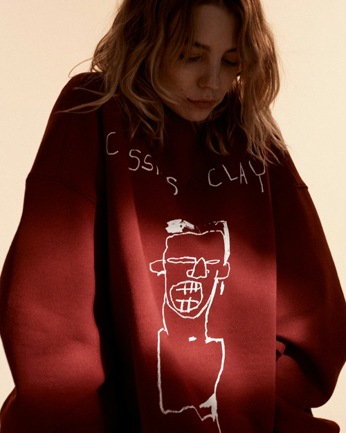 Young woman in a maroon sweatshirt featuring Basquiat-style artwork, gazing downward in a soft, diffused lighting setting.