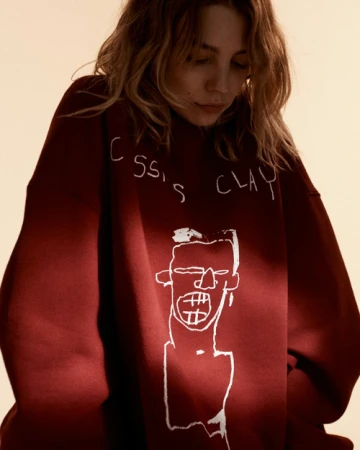 Young woman in a maroon sweatshirt featuring Basquiat-style artwork, gazing downward in a soft, diffused lighting setting.