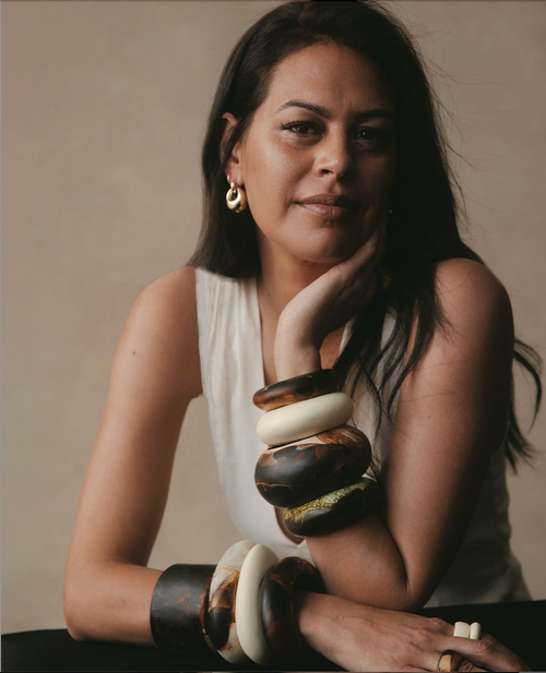 Portrait of a woman wearing a sleeveless top and multiple brown and white bangles, with her hand near her face, in a studio setting.
