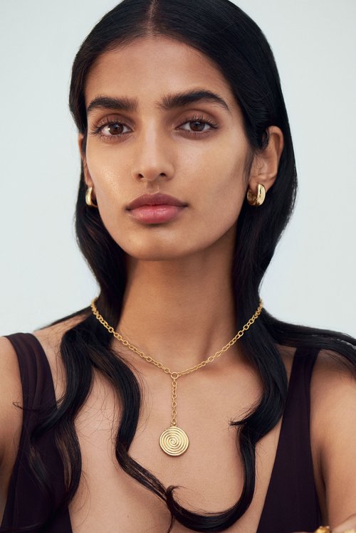 Close-up portrait of a woman with dark wavy hair and tan skin wearing a gold necklace and earrings against a light background.