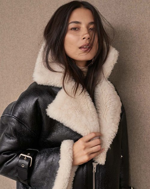 Portrait of a young adult woman wearing a black leather aviator jacket with cream shearling collar, standing indoors against a neutral backdrop.