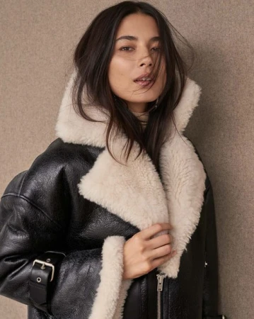 Portrait of a young adult woman wearing a black leather aviator jacket with cream shearling collar, standing indoors against a neutral backdrop.