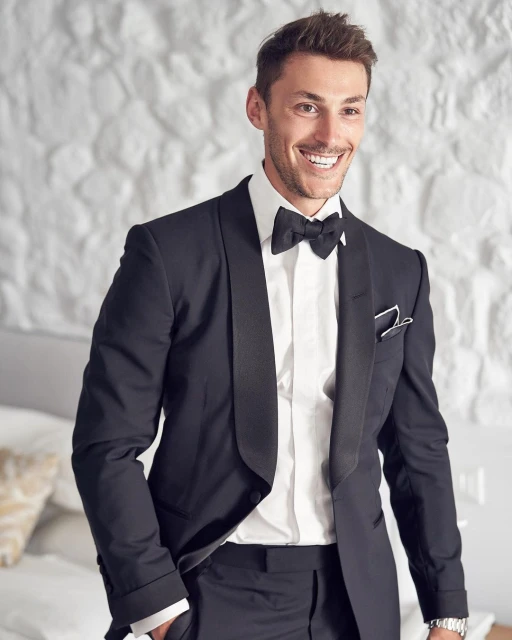 Smiling man in a black tuxedo and bow tie stands indoors against a textured white wall, looking confidently into the camera.