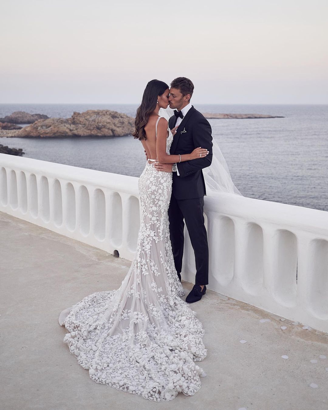 A wedding portrait captures a bride in a floral gown and groom in a black tuxedo kissing on a white balcony overlooking the ocean.