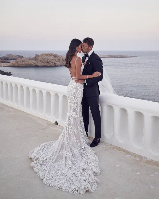 A wedding portrait captures a bride in a floral gown and groom in a black tuxedo kissing on a white balcony overlooking the ocean.