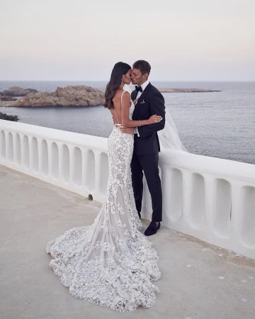 A wedding portrait captures a bride in a floral gown and groom in a black tuxedo kissing on a white balcony overlooking the ocean.