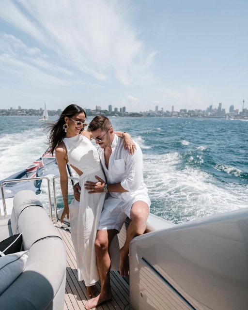 A stylish couple on a boat enjoys the sun, wearing white outfits and sunglasses, with a cityscape and blue water in the background.