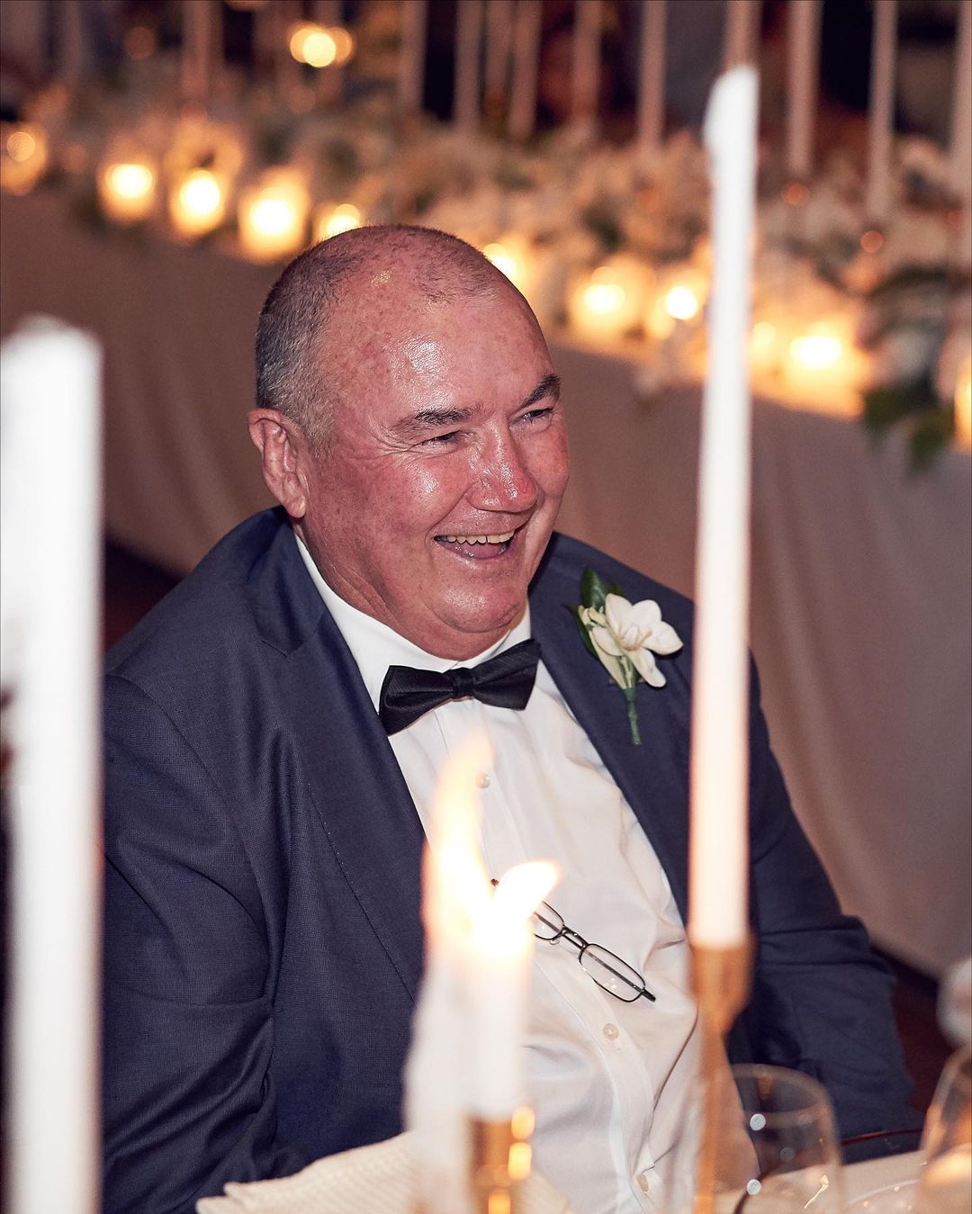 Smiling mature man in a suit with a bowtie sitting at an event table decorated with floral arrangements and candles, indoors.