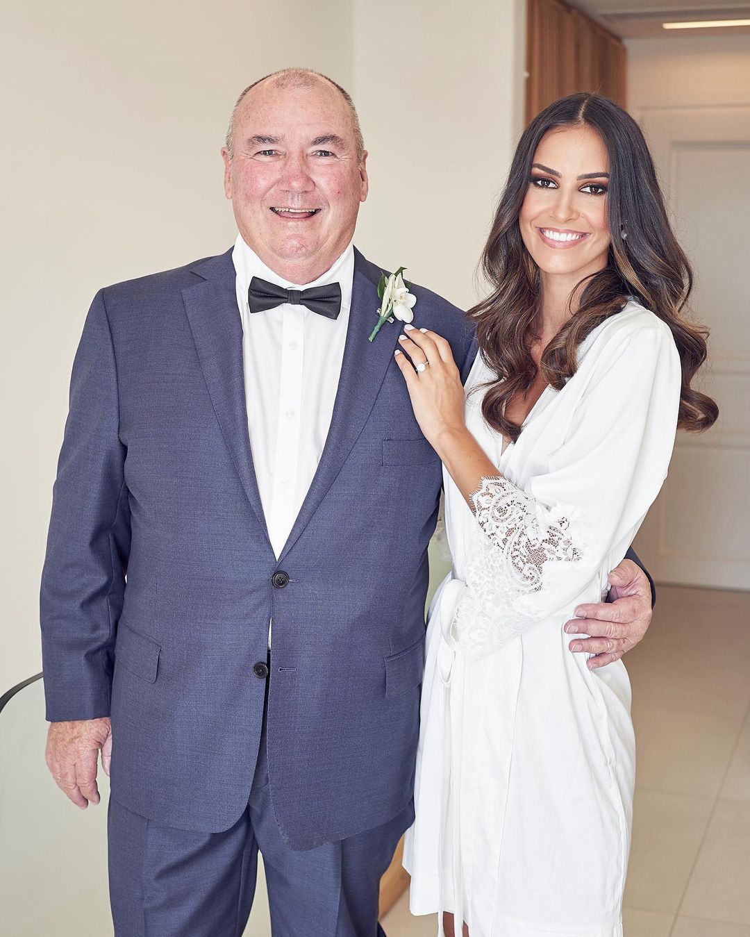 Father and daughter in an indoor setting, the father in a suit and the daughter in a white robe with lace sleeves. 