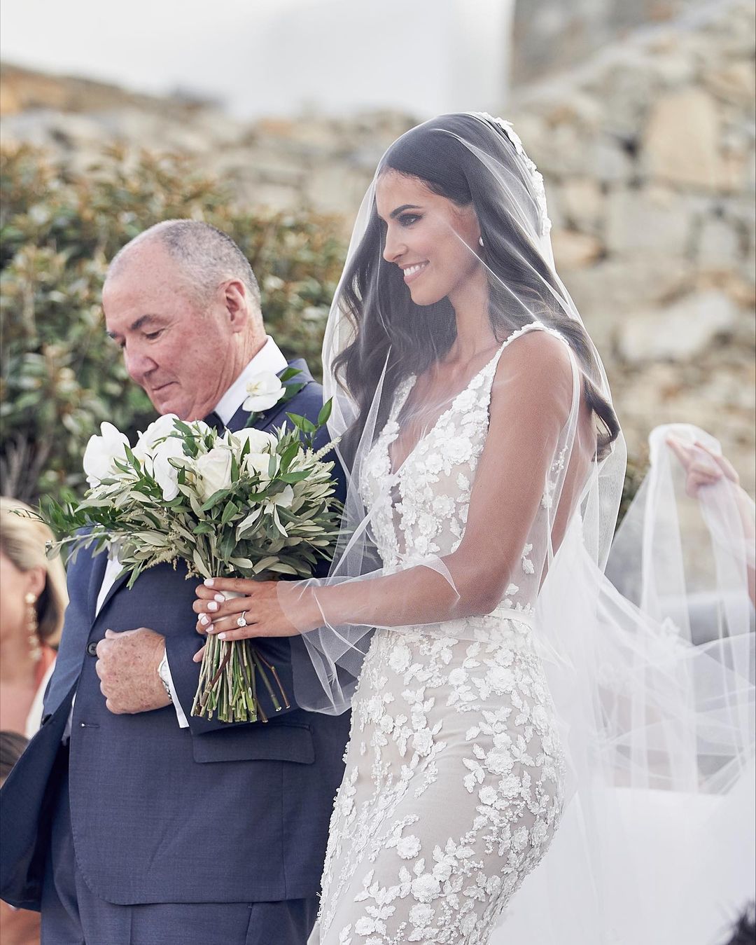 A bride in a floral lace dress with a long veil walks with an adult male holding a white rose bouquet in an outdoor setting.