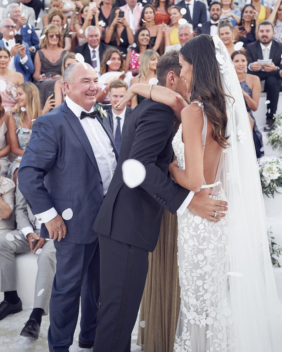 Wedding celebration with a bride and groom embracing, flower petals falling, and a crowd of guests in the background on a sunny day.
