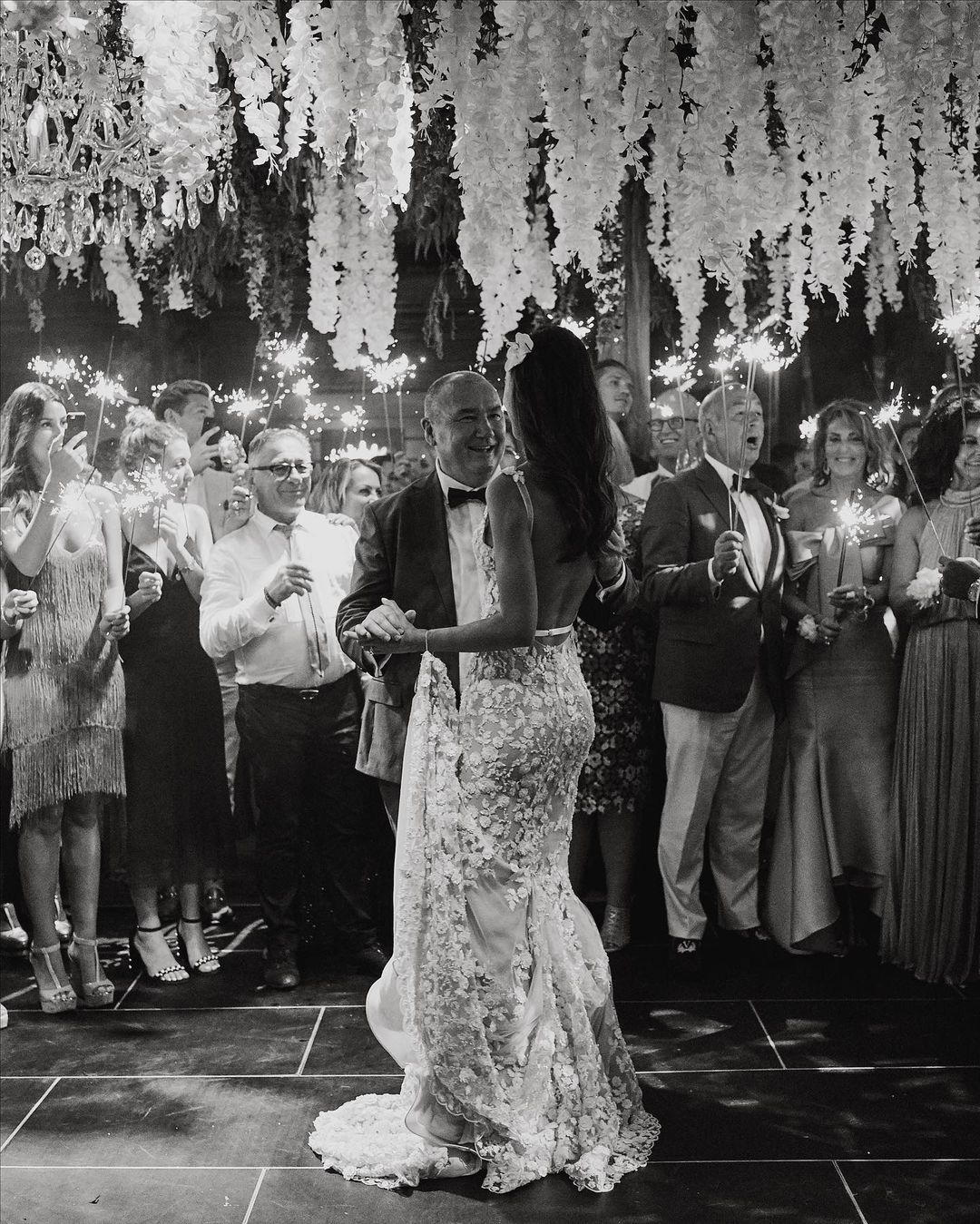 Monochrome wedding reception scene with bride and groom dancing surrounded by guests holding sparklers under floral decorations.