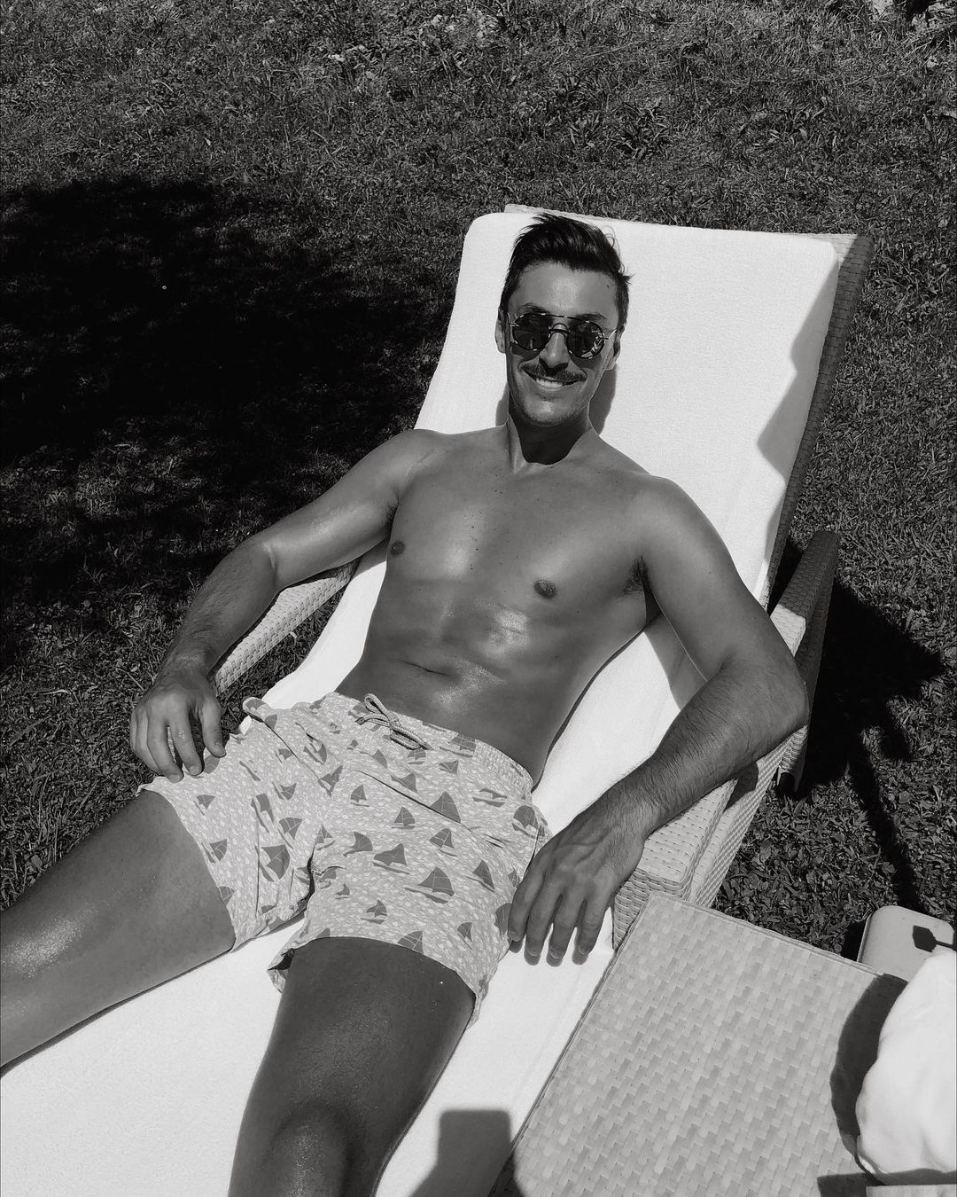 Monochrome portrait of a shirtless adult male with a mustache and sunglasses, lying on a white lounge chair outdoors in the sunshine.