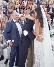 Bride in a white lace gown hugging a groom in a black suit with guests watching as white petals fall around them.