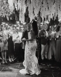 Bride and groom dancing at a wedding reception surrounded by guests holding lit sparklers under hanging floral decor
